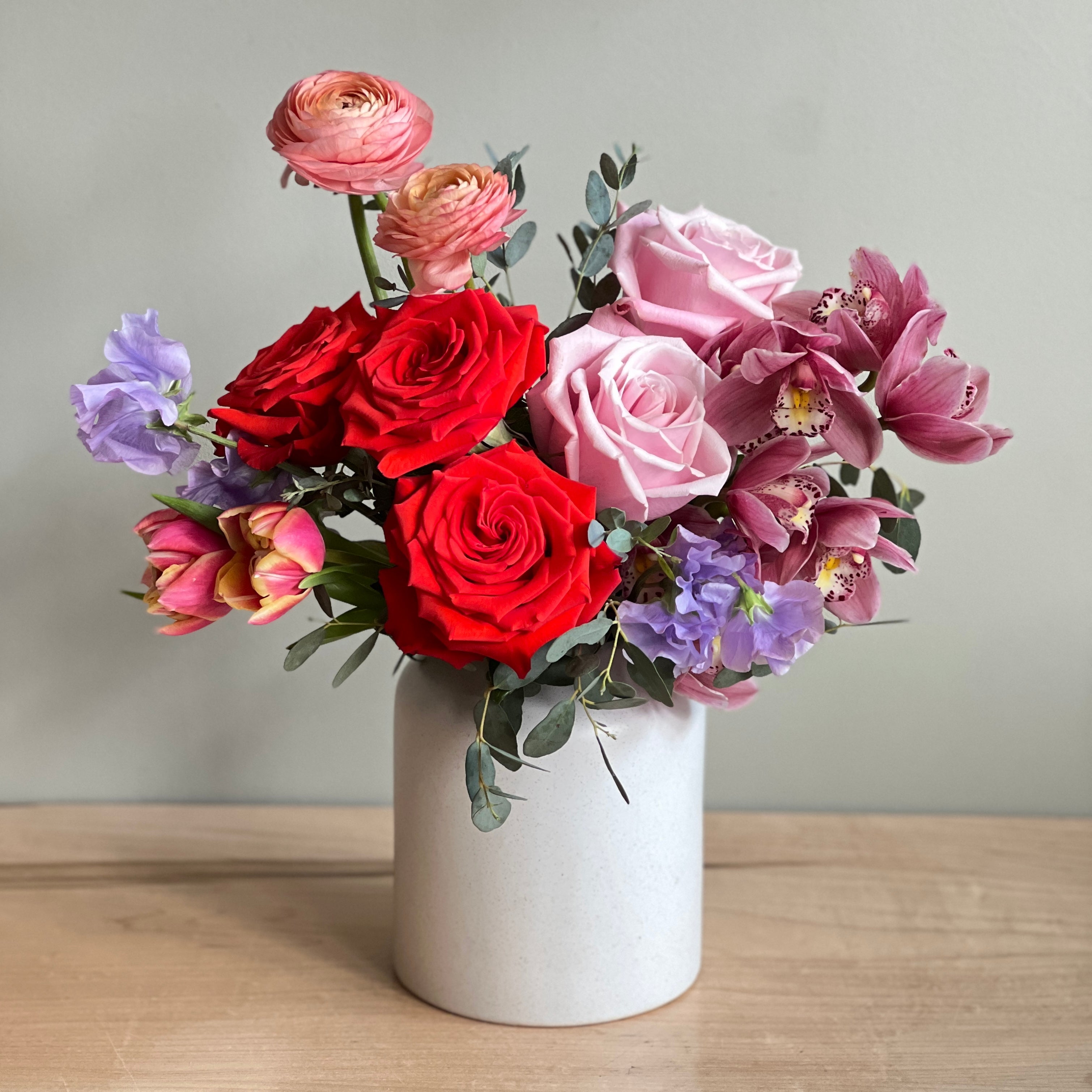 Flower arrangement in bright red, peach, light pink, and lavender in a beige ceramic vase on a wood table.