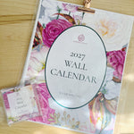 A floral 2027 wall calendar featuring twelve illustrations by local artist, Wickie Roland. Pictured n a clear plastic sleeve with a rose gold clip, next to a smaller matching mini calendar.