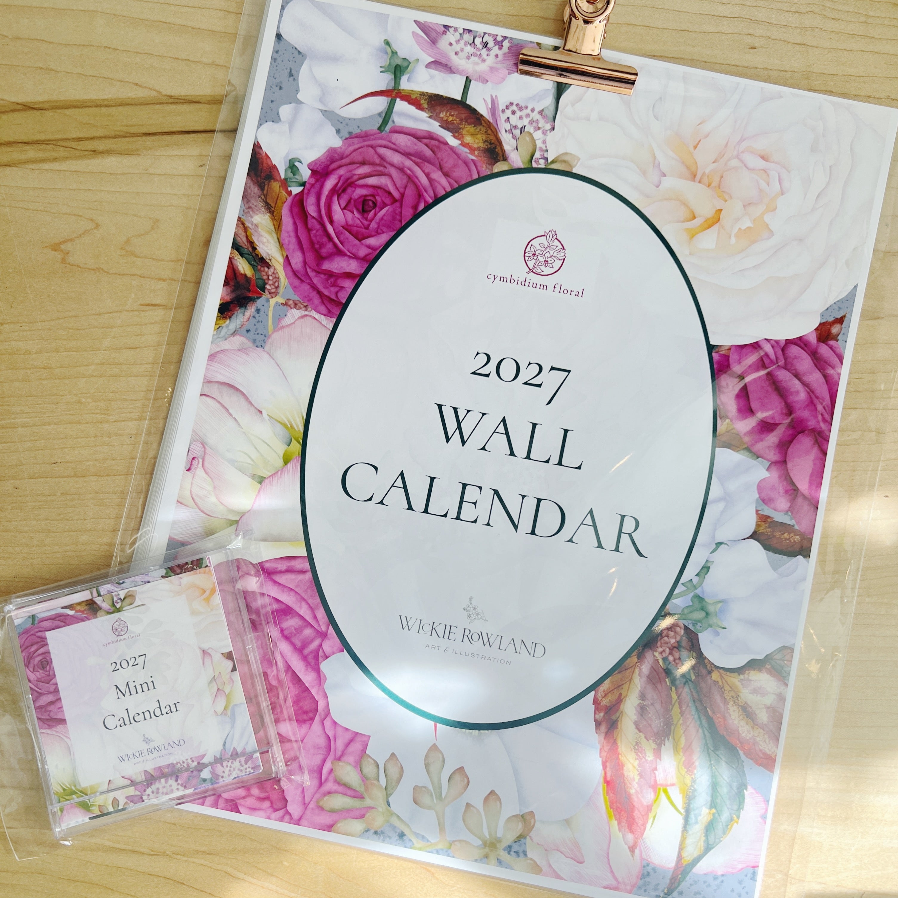 A floral 2027 wall calendar featuring twelve illustrations by local artist, Wickie Roland. Pictured n a clear plastic sleeve with a rose gold clip, next to a smaller matching mini calendar.