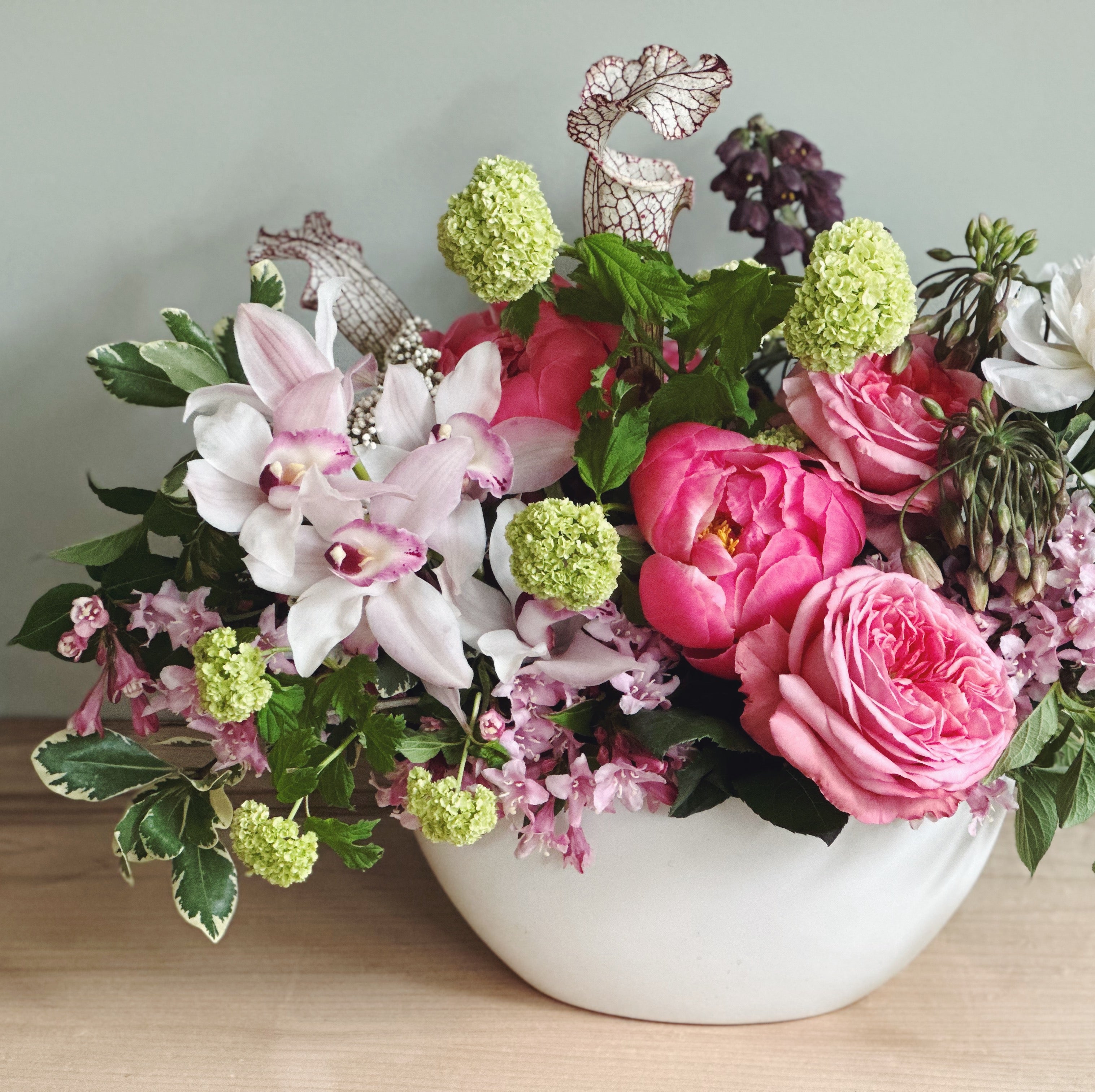 High-end, luxe bouquet of premium flowers featuring peonies, roses, orchids and locally grown natural textures in a white vase on a wooden surface with a light gray background