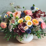 High-end, luxe colorful flower arrangement with premium orchids, roses, dahlias, ranunculus and locally grown natural textures in a white vase on a wooden surface with a light gray background