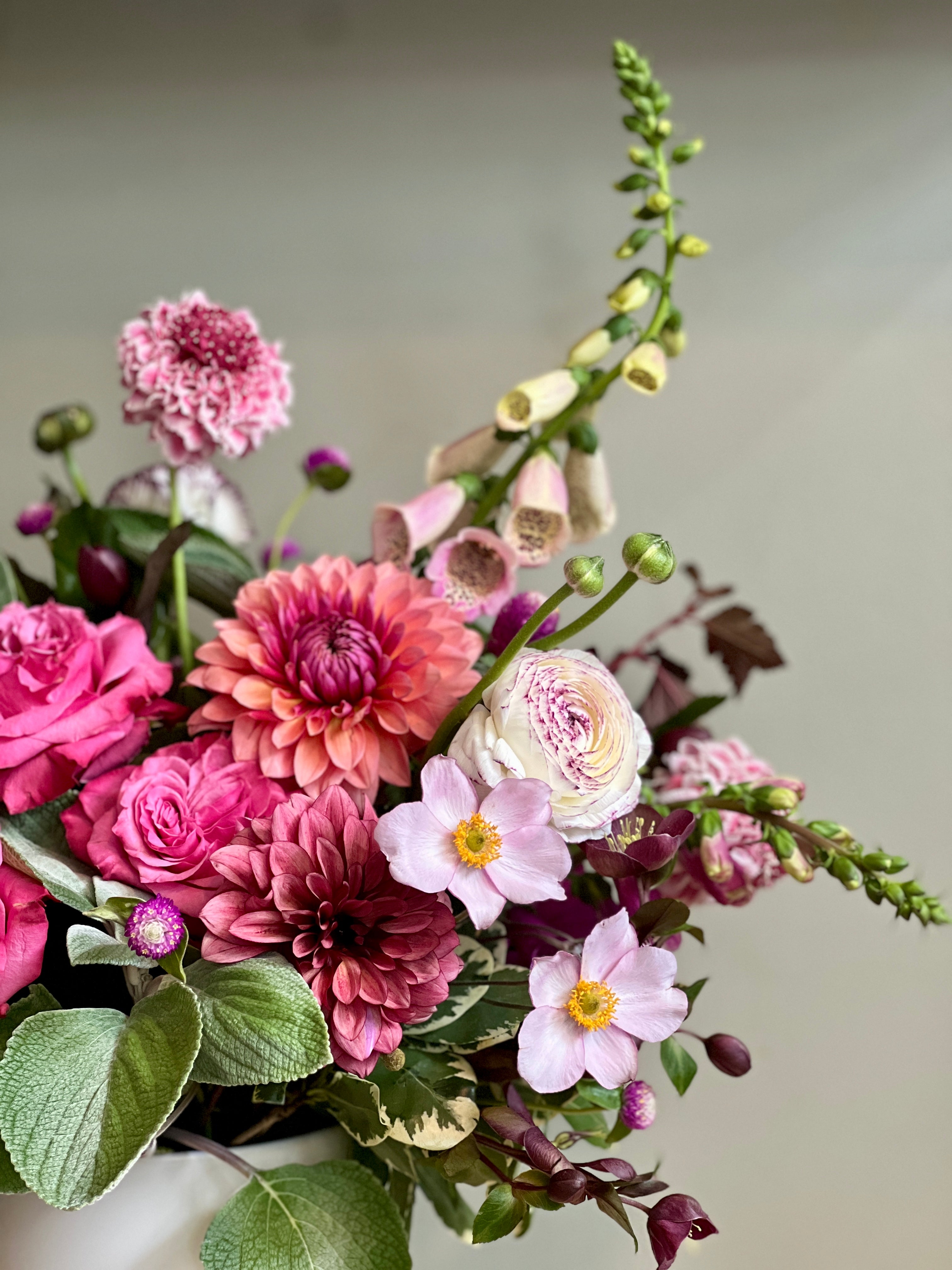 A vibrant floral arrangement featuring bright pink and magenta roses, pink dahlias, light pink daisies, and green foliage, set against a neutral light gray background.