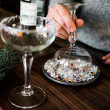 Close-up of a hand holding a glass over a dish with floral salt on a wooden surface.