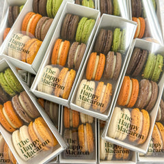 Several white rectangular boxes filled with assorted macarons arranged on a metallic gray surface. The macarons include orange, green, brown, beige, and cream colors, and the boxes are printed with the text 'The Happy Macaron'.