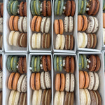 Multiple white rectangular boxes filled with assorted French macarons arranged in neat rows. The macarons come in various colors including orange, brown, green, cream, and speckled beige.