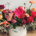 Colorful fall colored flower arrangement with amaryllis, orchids and ranunculus in a white vase on a wooden surface with a neutral background