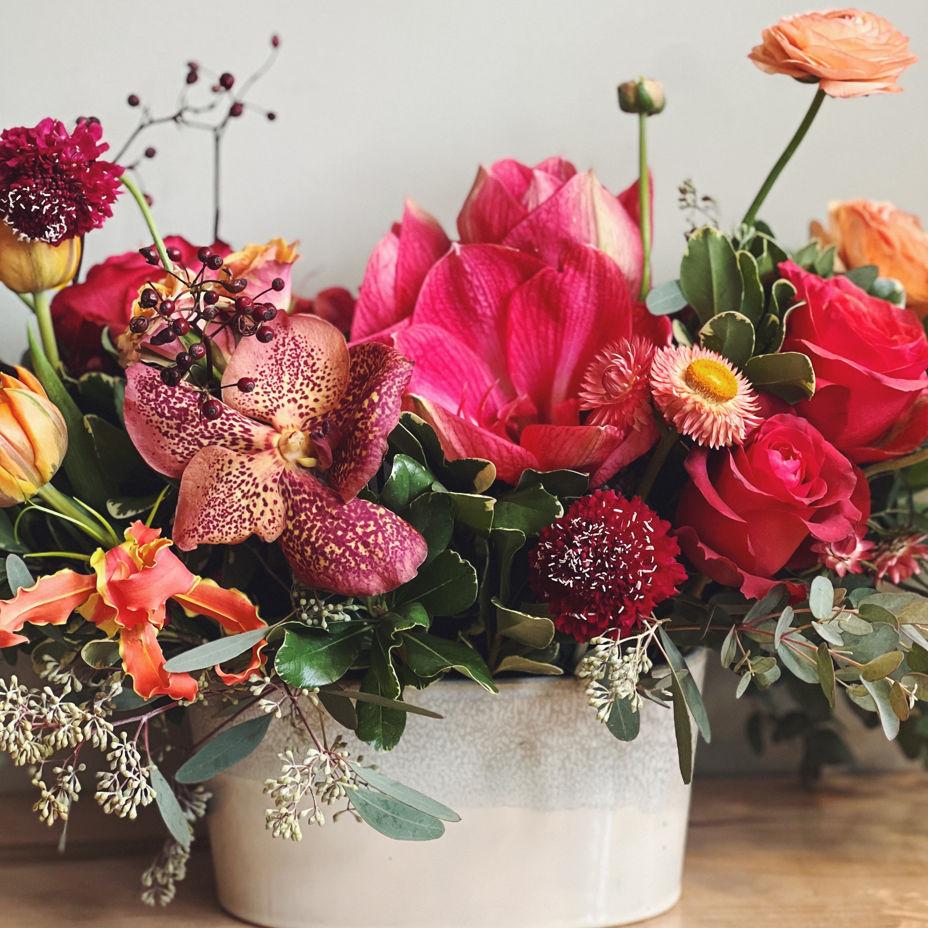 Colorful fall colored flower arrangement with amaryllis, orchids and ranunculus in a white vase on a wooden surface with a neutral background