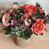 Bouquet of rust and burgundy with roses and orchids in a vase on a wooden surface.