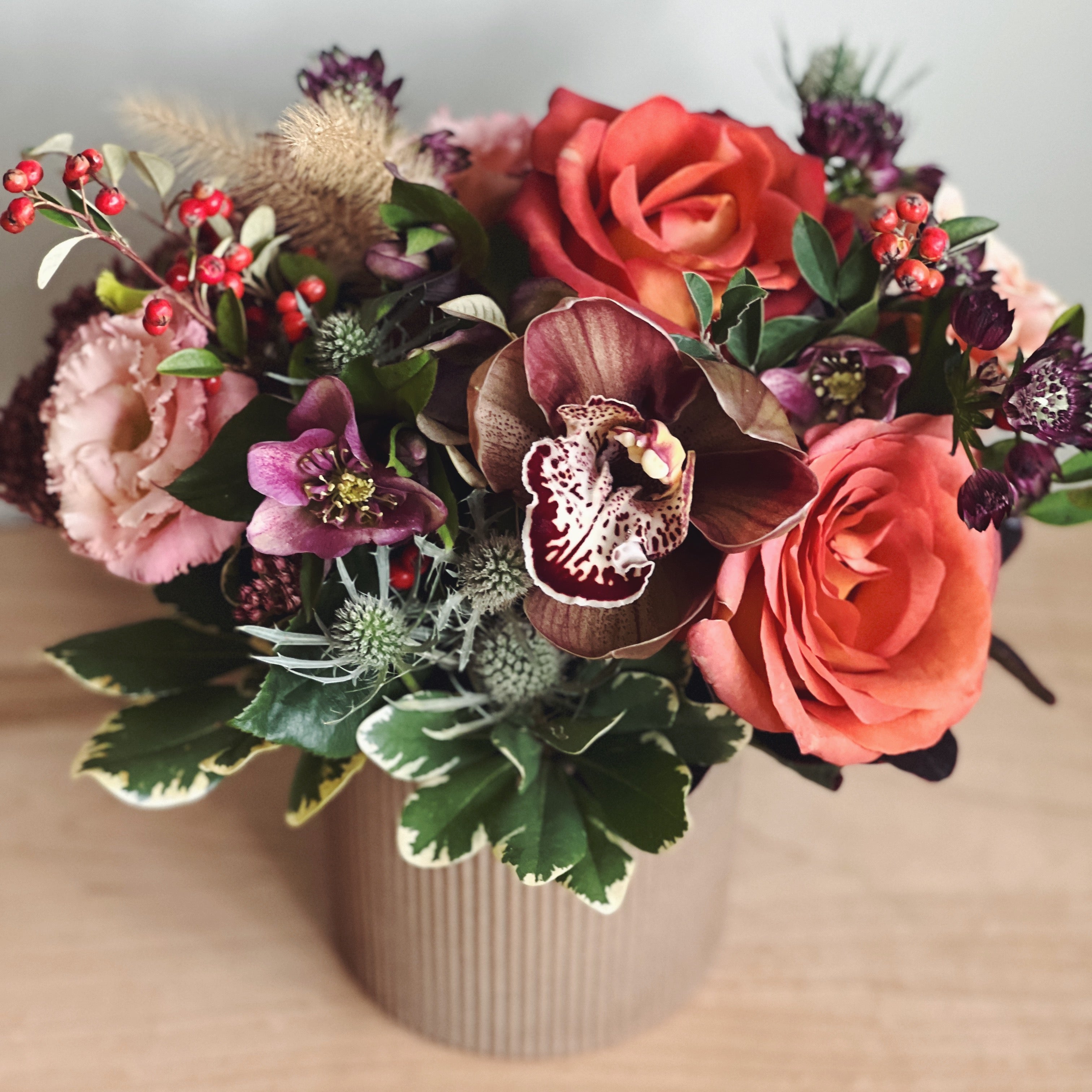 Bouquet of rust and burgundy with roses and orchids in a vase on a wooden surface.