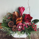 Holiday floral arrangement with burgundy orchids, brown and green magnolia leaves, pinecones, and evergreens in a white ridged oval ceramic container.