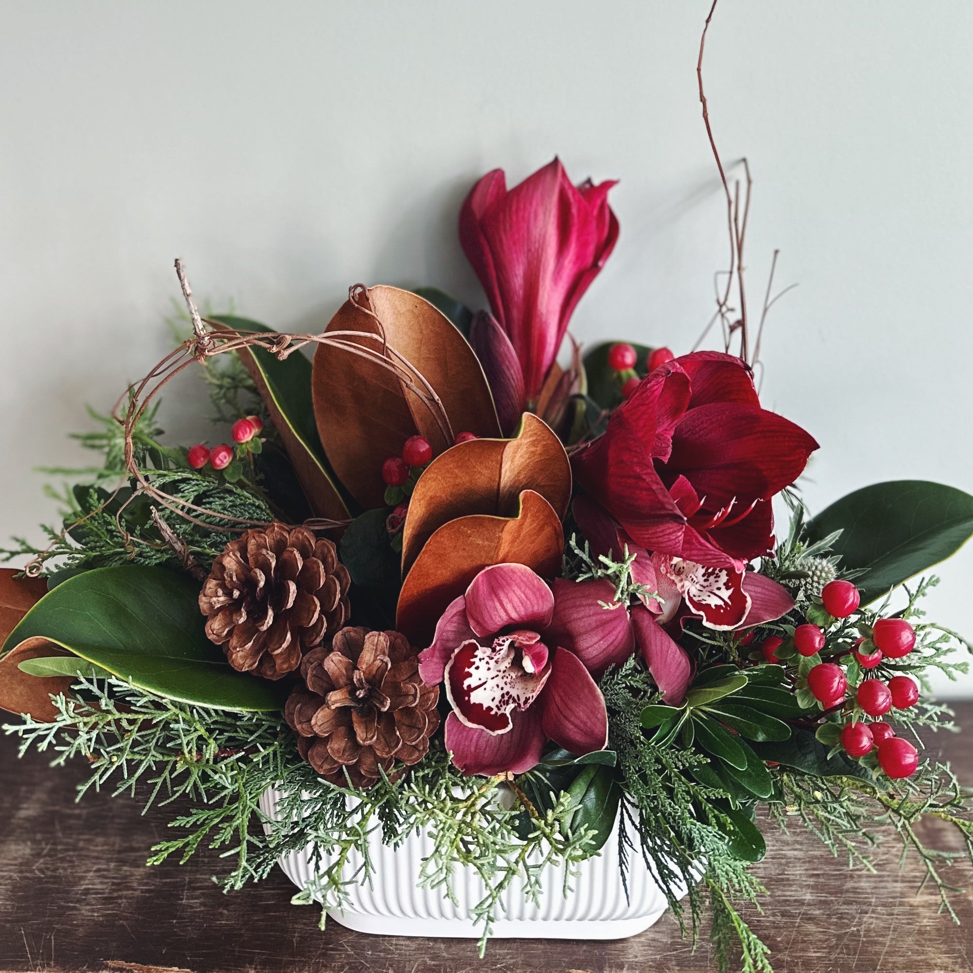 Holiday floral arrangement with burgundy orchids, brown and green magnolia leaves, pinecones, and evergreens in a white ridged oval ceramic container.