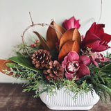 Holiday floral arrangement with burgundy orchids, brown and green magnolia leaves, pinecones, and evergreens in a white ridged oval ceramic container.