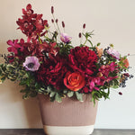 Burgundy, magenta, lavender, and deep red holiday luxe flower arrangement in a beige oval ceramic vase.  Orchids, anemones, peonies, roses, sanguisorba, and privet berries.