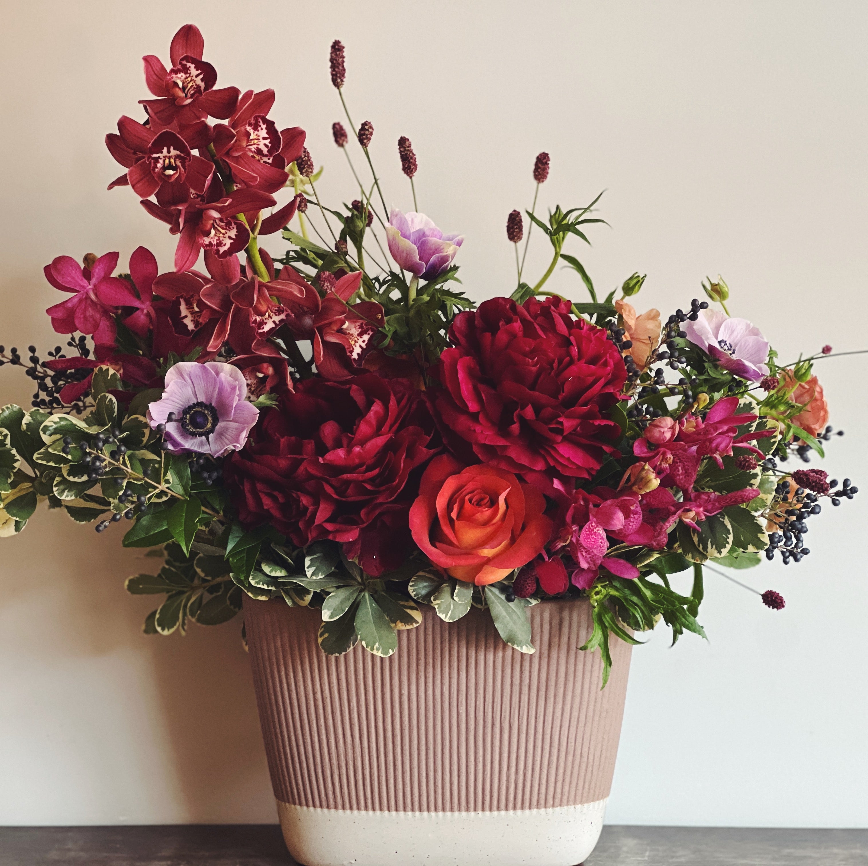 Burgundy, magenta, lavender, and deep red holiday luxe flower arrangement in a beige oval ceramic vase.  Orchids, anemones, peonies, roses, sanguisorba, and privet berries.