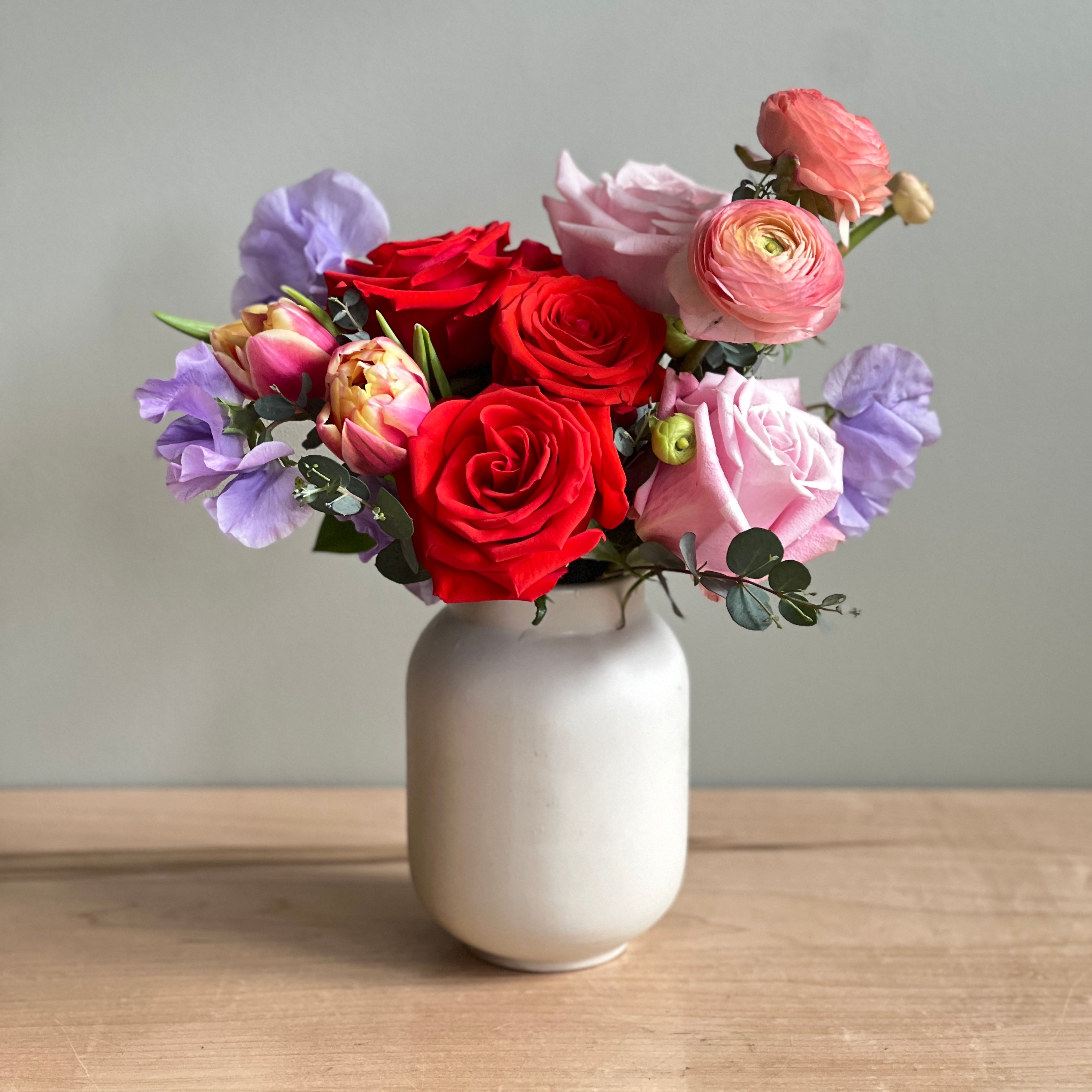 Unique Valentine's Day flowers in a ceramic vase featuring roses, ranunculus, tulips, and sweetpea in shades of pinks, red and lavender. 