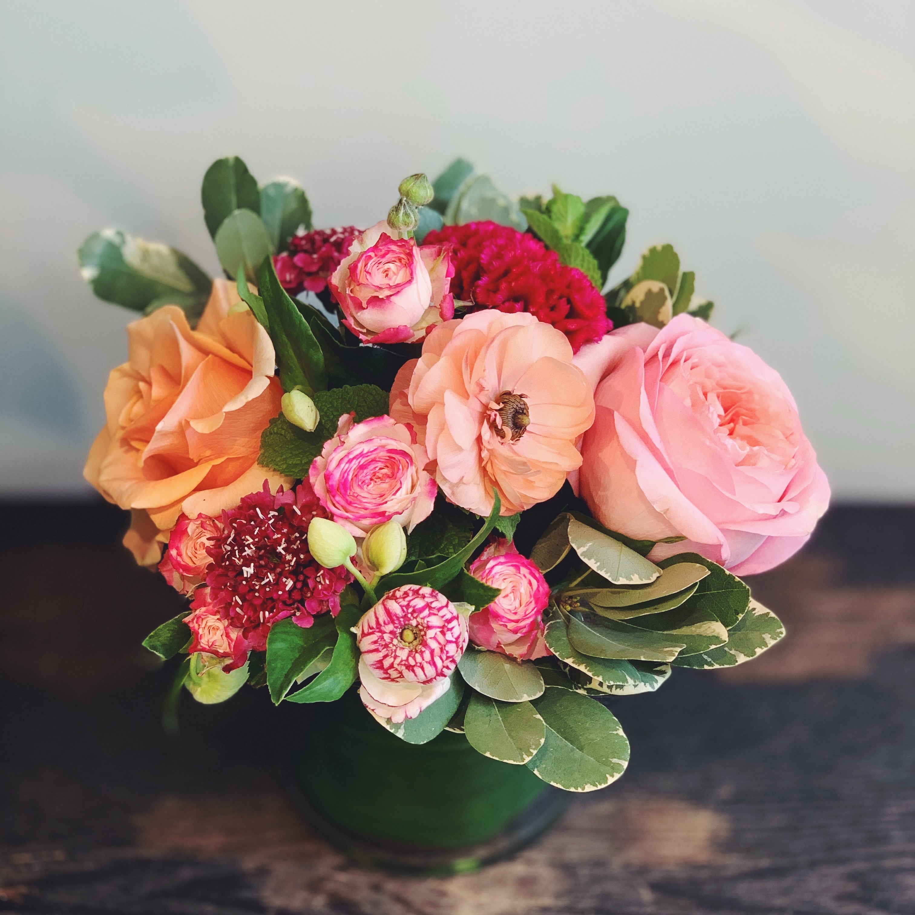 Bold and bright lush and low arrangement in orange, peach,  raspberry, and hot pink.  Flowers include garden-like roses, scabiosa, celoisa, and mint.