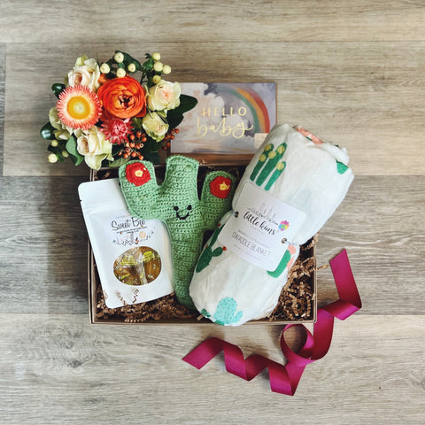Kraft colored cardboard gift box filled with kraft colored crinkle.  Gift items nested into the box include a mini flower arrangement, green cactus shaped rattle, a baby swaddle blanket white with various cacti, a bag of honey candies, and a Greeting card in matching colors.