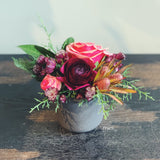 Mini holiday flower arrangement in plum, coral, and brown.  Flowers include: roses, astrantia, ranunculus, and leucadendron accented by evergreens.