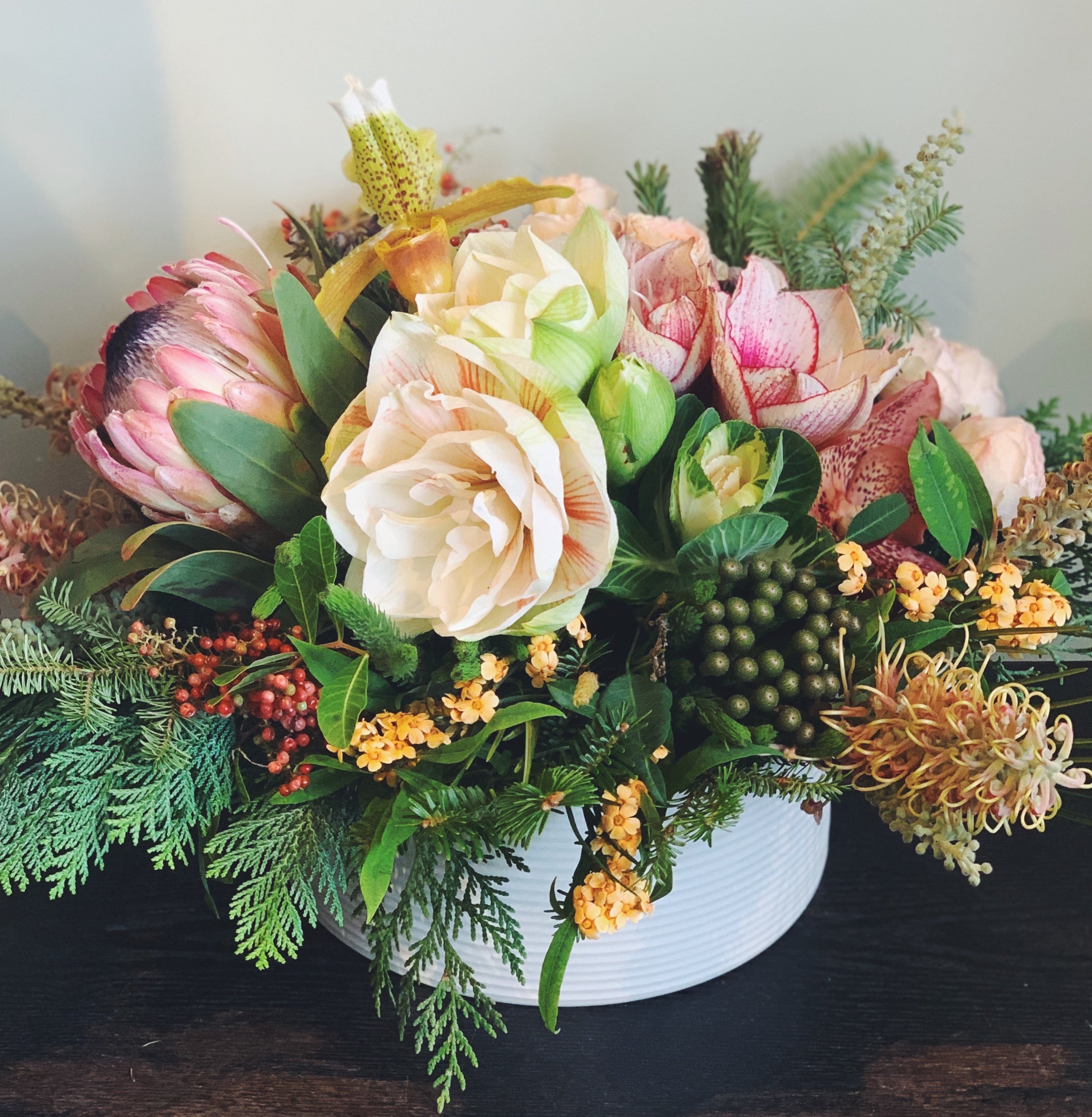 Holiday flower arrangement in pale pink, cream, green, and red. Flowers include: amaryllis, pink protea, lady slipper orchid, kale, pepperberry, and evergreens.