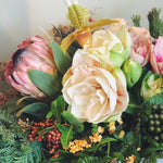 Holiday flower arrangement in pale pink, cream, green, and red.  Flowers include: amaryllis, pink protea, lady slipper orchid, kale, pepperberry, and evergreens. 